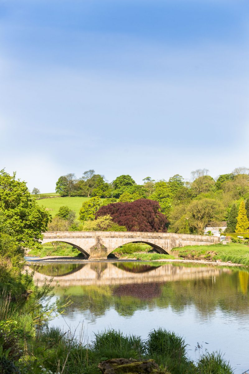A bridge spannijng a wide river - surrounded by lush green trees and fields